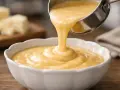 Une louche verse une sauce fromagère onctueuse dans un bol blanc. Des blocs de fromage sont flous en arrière-plan.