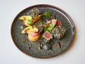 Un délicieux tataki de thon enrobé de sésame, servi avec de l'avocat, des oignons rouges et une sauce onctueuse sur une assiette texturée.