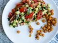 Salade colorée de tomates, concombres et pois chiches, garnie de petites fleurs blanches. Des pois chiches rôtis sont disposés à côté.