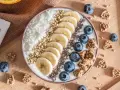Un bol de smoothie garni de bananes, myrtilles, noix de coco râpée et graines. Une recette pas cher et saine pour un petit-déjeuner gourmand.
