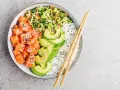 Un délicieux base poke bowl avec saumon cru, avocat, riz blanc et légumes croquants, prêt à être dégusté avec des baguettes.