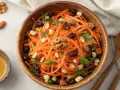 Salade de carottes râpées, garnie de noix, raisins secs et persil, dans un bol en bois. Un petit ramequin de miel est à côté.