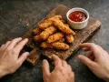 Des doigts s'approchent d'un tas de poulet pané croustillant sur une planche en bois, avec un petit ramequin de sauce tomate.