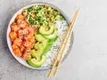 Un délicieux poke bowl santé avec saumon cru, avocat, riz blanc et légumes croquants, prêt à être dégusté avec des baguettes.