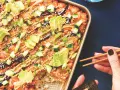 Un délicieux sushi bake dans un plat doré, garni d'avocat, concombre, oignons verts et graines de sésame. Une main tient des baguettes pour servir.