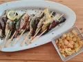 Sardines grillées sur brochettes, garnies de citron et persil, accompagnées de pommes de terre sautées dans un panier métallique.