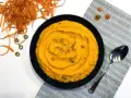 Un bol de purée de carotte onctueuse, garnie de graines, à côté d'une cuillère et de carottes râpées.
