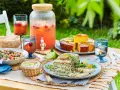 Un délicieux repas d'été entre amis : salade, terrine, gâteau, et une grande carafe de boisson fruitée, le tout sur une table dressée en extérieur.
