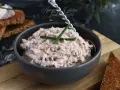 Un bol de rillettes de thon crémeuses, garni de ciboule, prêt à être tartiné sur des toasts grillés.