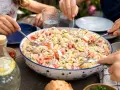 Un grand plat de salade de pâtes au surimi, tomates et thon, servi à l'extérieur. Des mains servent la salade.