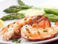 Queue de homard grillée, servie avec des asperges et une sauce homard crémeuse, garnie de quartiers de citron et de thym frais.