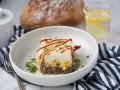 Un délicieux pâté chinois servi dans une assiette blanche, garni de persil et de ketchup. Un verre d'eau citronnée et du pain complètent ce plat réconfortant.