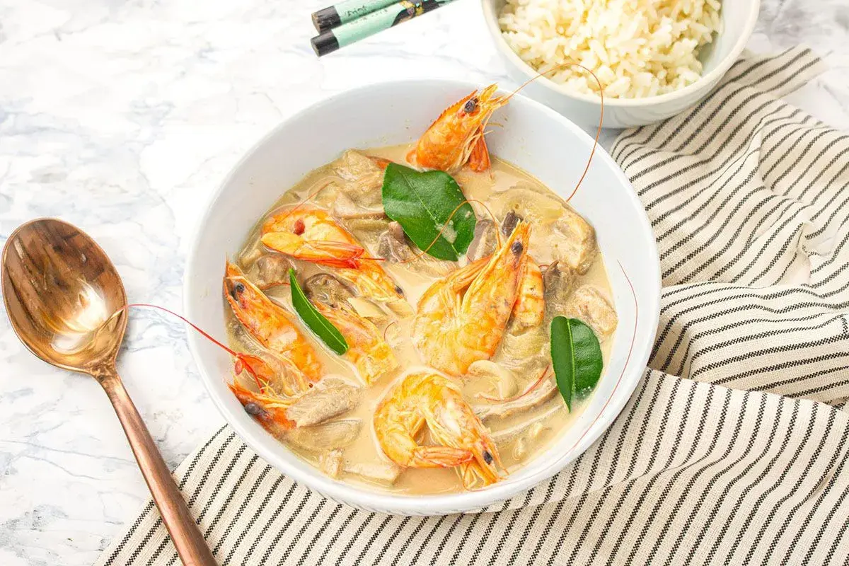 Un plat de sauté crevettes thaï lait de coco, garni de feuilles vertes, servi avec du riz et une cuillère en cuivre.