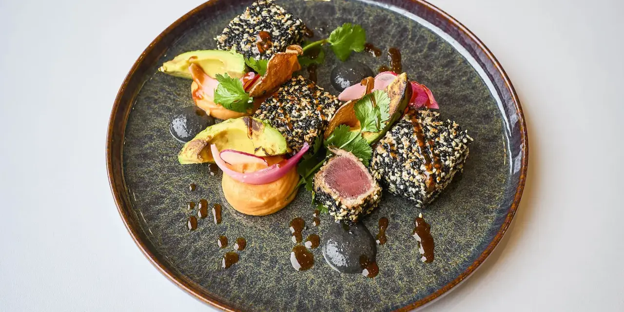 Un délicieux tataki de thon enrobé de sésame, servi avec de l'avocat, des oignons rouges et une sauce onctueuse sur une assiette texturée.