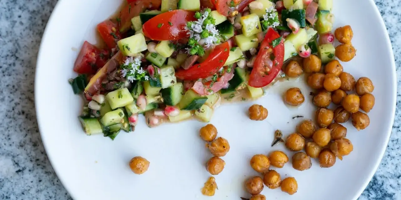Salade colorée de tomates, concombres et pois chiches, garnie de petites fleurs blanches. Des pois chiches rôtis sont disposés à côté.