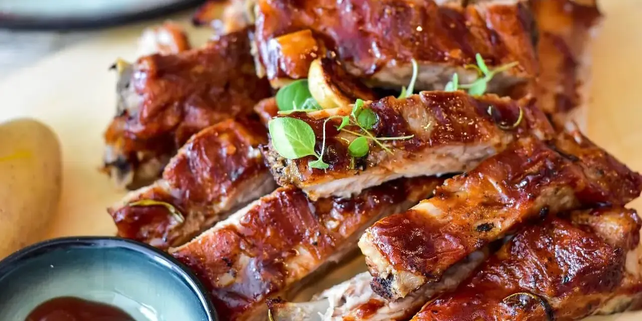 Délicieux ribs au four, nappés d'une sauce barbecue brillante, servis avec une petite coupelle de sauce et des herbes fraîches.