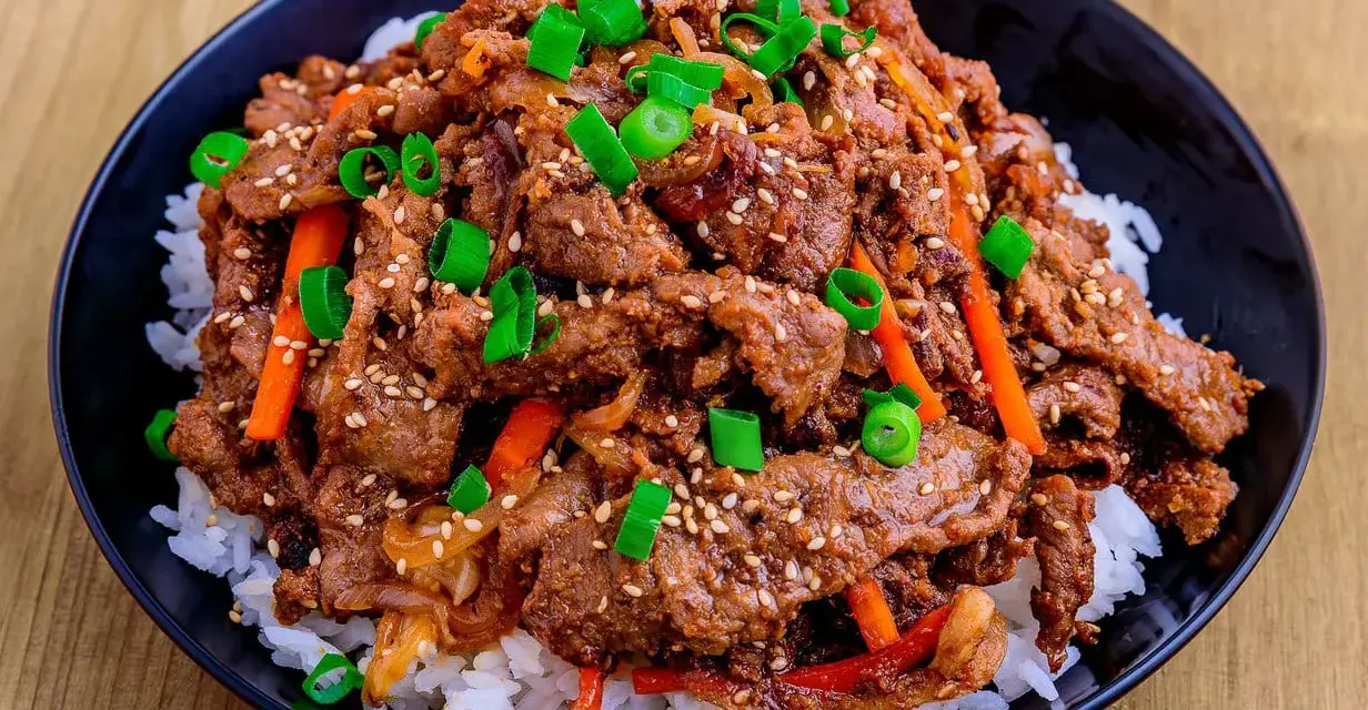Un bol de riz blanc garni de boeuf bulgogi mariné, de julienne de carottes et d'oignons verts ciselés, parsemé de graines de sésame.