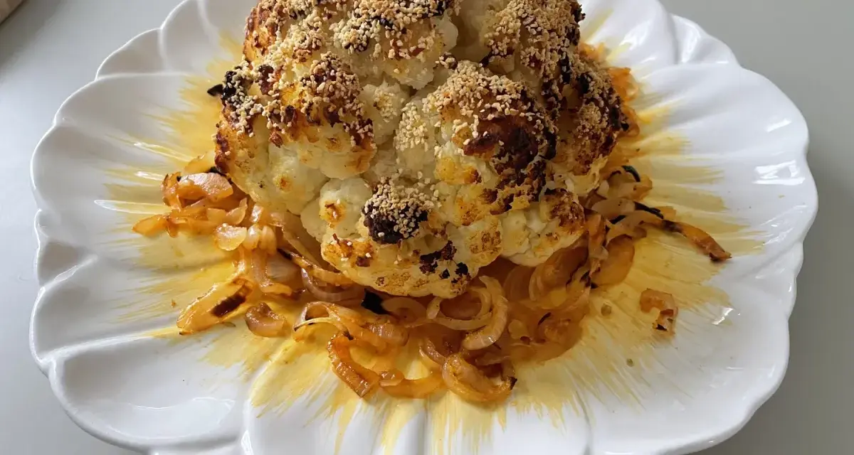 Un chou-fleur entier, rôti au four, est servi sur une assiette blanche ornée d'oignons caramélisés. Le chou-fleur est saupoudré de graines de sésame et d'épices.