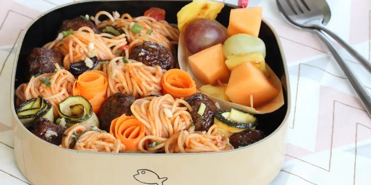 Bento box avec spaghetti et boulettes, garni de courgettes et carottes roulées. Une brochette de fruits complète cette idée repas à emporter au travail.