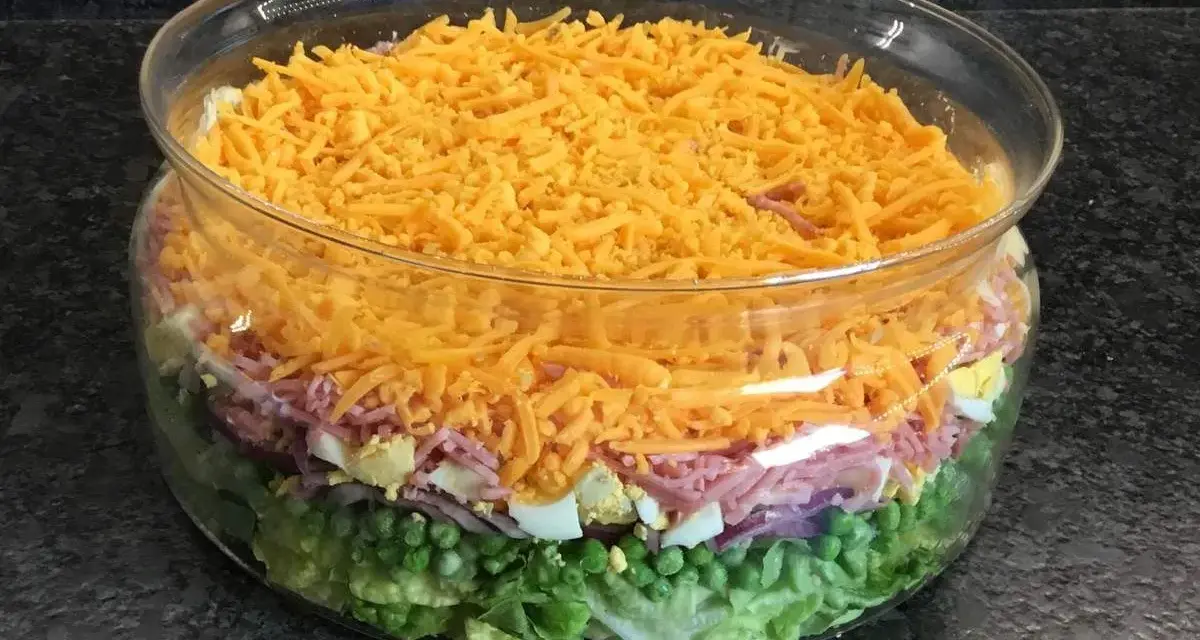 Salade à étage colorée dans un bol en verre : laitue, petits pois, oignons rouges, jambon, œufs durs et fromage râpé.
