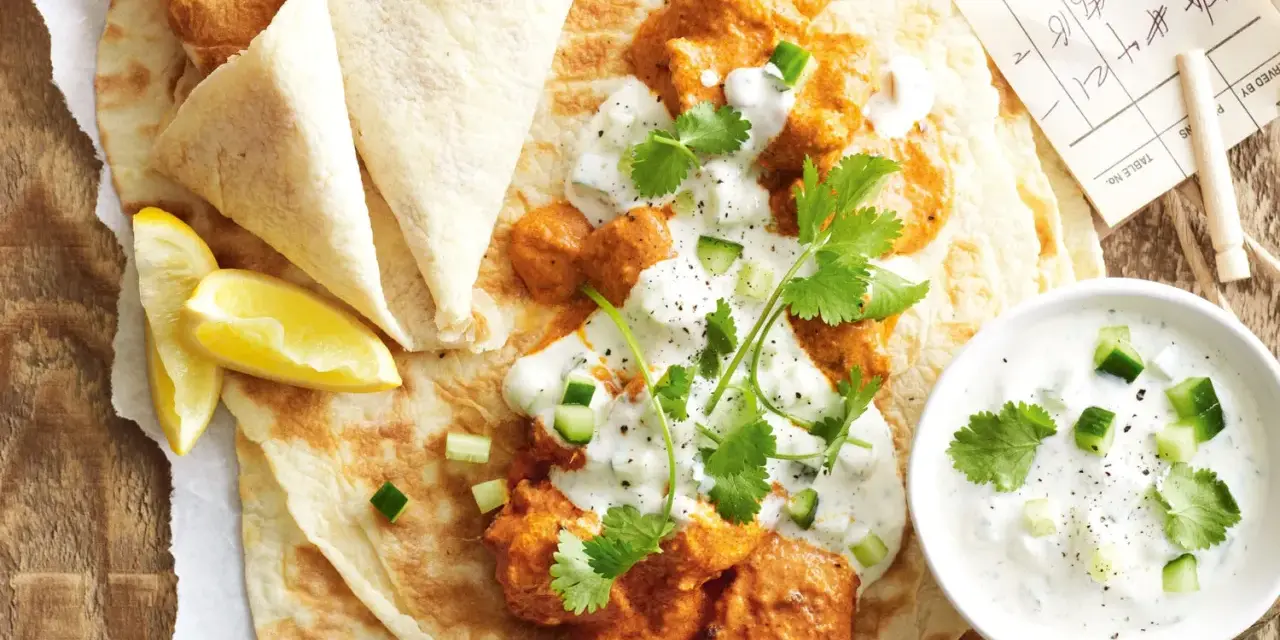Un plat de butter chicken crémeux servi sur du pain naan avec du raïta et des quartiers de citron.