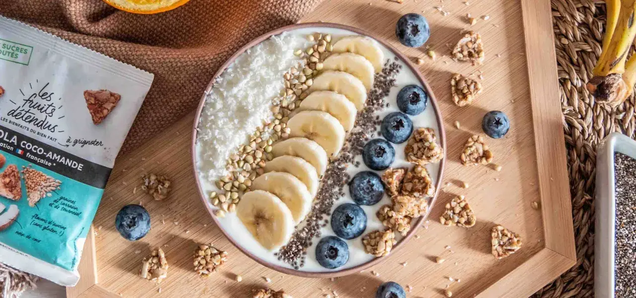 Un bol de smoothie garni de bananes, myrtilles, noix de coco râpée et graines. Une recette pas cher et saine pour un petit-déjeuner gourmand.