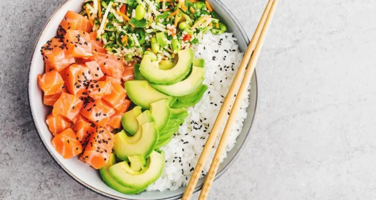Un délicieux base poke bowl avec saumon cru, avocat, riz blanc et légumes croquants, prêt à être dégusté avec des baguettes.