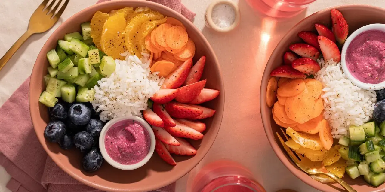 Deux bols colorés remplis de riz, fraises, myrtilles, concombre, carottes et oranges. Une belle idée poke bowl pour un repas frais et sain.