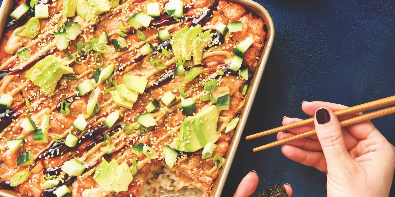 Un délicieux sushi bake dans un plat doré, garni d'avocat, concombre, oignons verts et graines de sésame. Une main tient des baguettes pour servir.