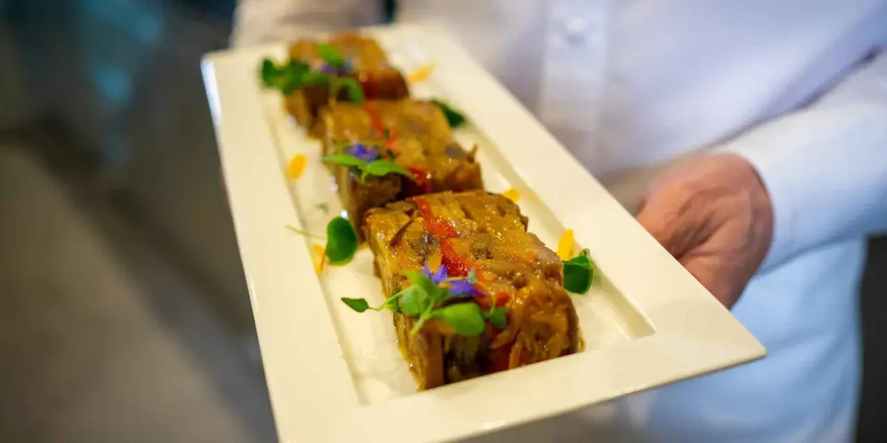 Trois tranches de terrine de légumes, garnies de fleurs comestibles et de verdure, présentées sur une assiette blanche par un serveur.
