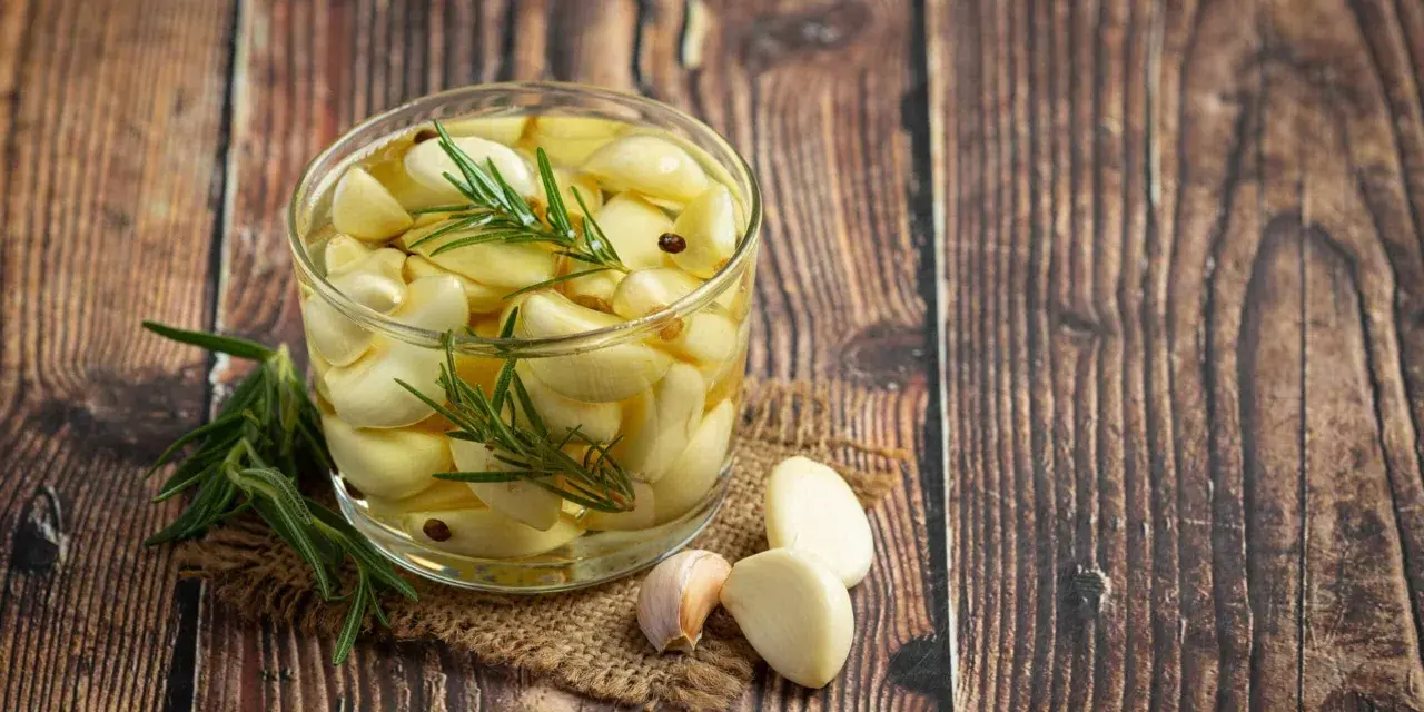 Un bocal de **ail confit** avec des brins de romarin et quelques grains de poivre, posé sur une toile de jute, sur un fond de bois rustique.