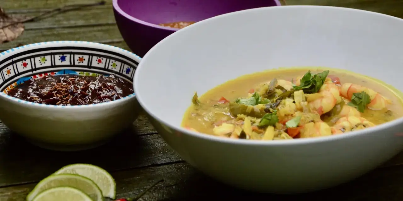 Un plat de crevette curry coco crémeux, garni de coriandre fraîche, servi avec des quartiers de citron vert et un piment rouge.