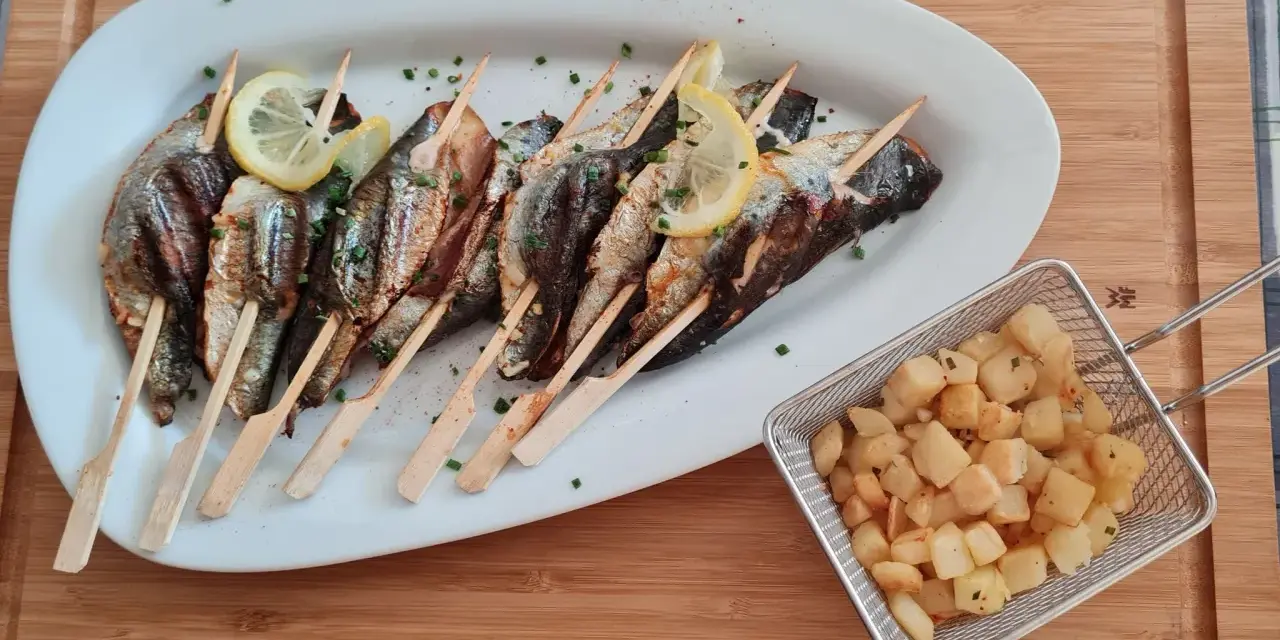 Sardines grillées sur brochettes, garnies de citron et persil, accompagnées de pommes de terre sautées dans un panier métallique.