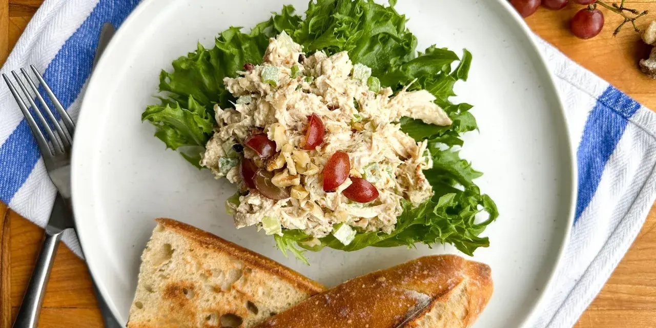Salade de poulet froid mayonnaise sur laitue, avec des raisins et des noix, servie avec du pain croustillant.