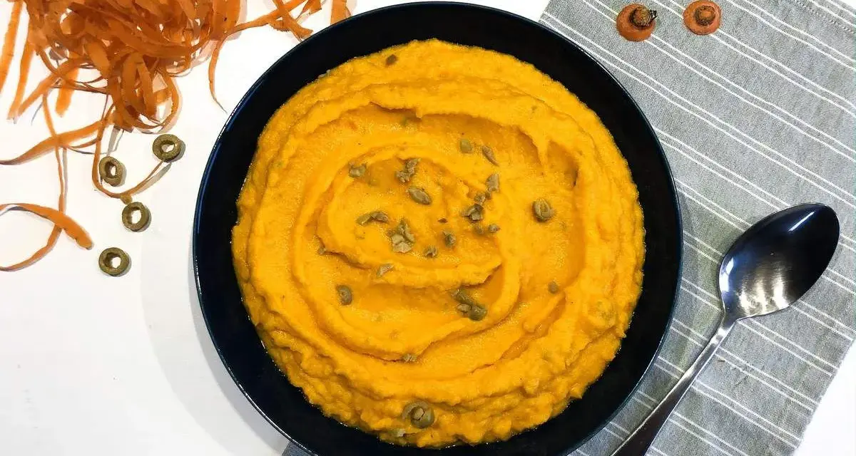 Un bol de purée de carotte onctueuse, garnie de graines, à côté d'une cuillère et de carottes râpées.