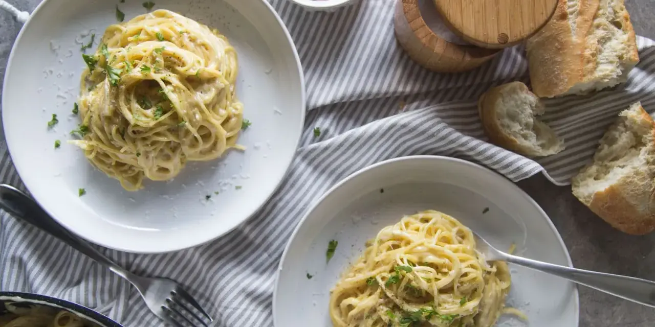 Deux assiettes de pâtes crémeuses à l'ail, garnies de persil frais. Un pain croustillant et du fromage râpé complètent cette recette de pâtes à l'ail et crème.
