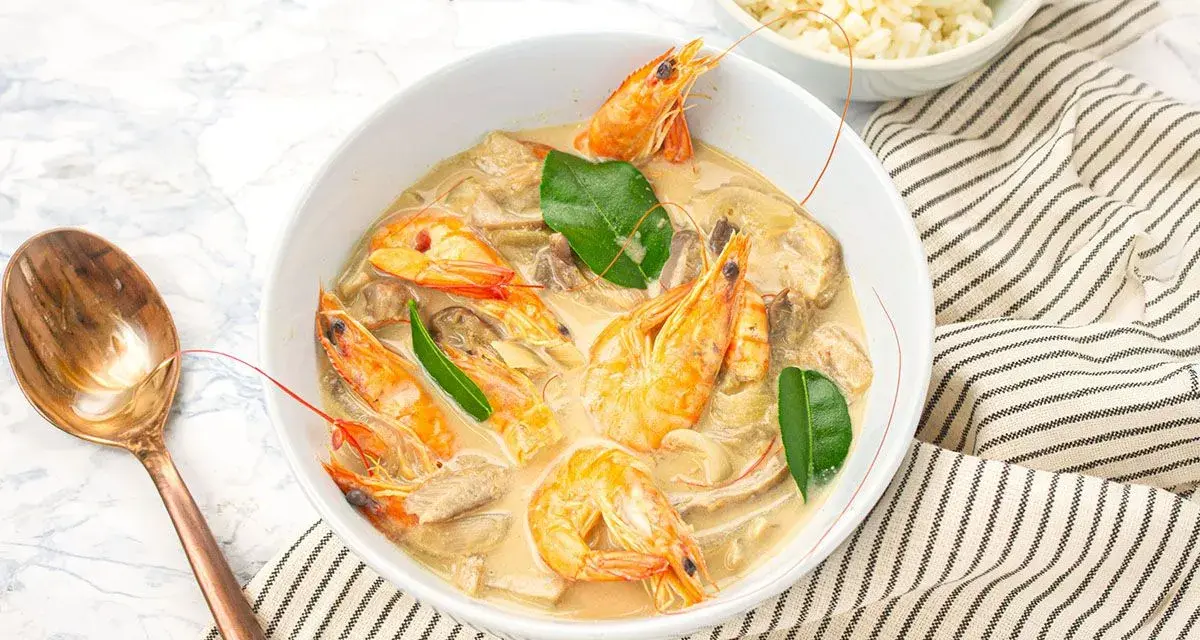 Un plat de sauté crevettes thaï lait de coco, garni de feuilles vertes, servi avec du riz et une cuillère en cuivre.