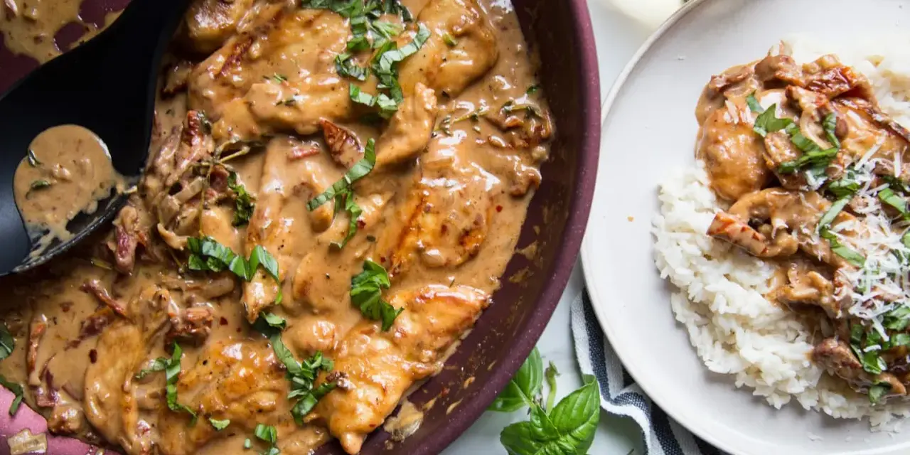 Poulet en sauce tomate séchée crème, garni de basilic frais, servi dans une poêle et sur une assiette de riz.