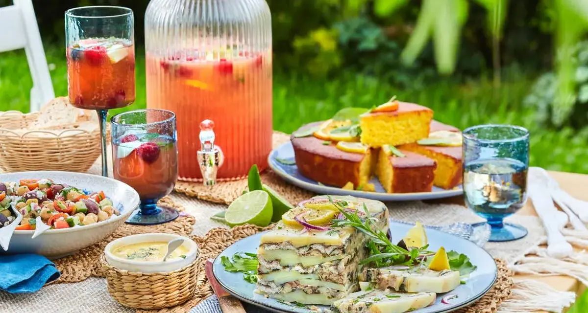 Un délicieux repas d'été entre amis : salade, terrine, gâteau, et une grande carafe de boisson fruitée, le tout sur une table dressée en extérieur.