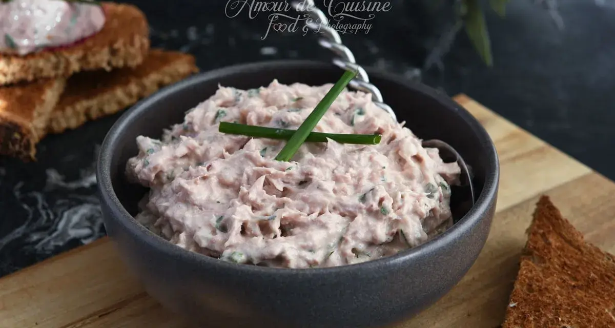 Un bol de rillettes de thon crémeuses, garni de ciboule, prêt à être tartiné sur des toasts grillés.