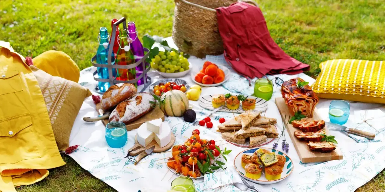 Un pique-nique gourmand sur l'herbe : pains, fromages, fruits, mini quiches, sandwichs, roulé salé, et boissons colorées. Une belle idée pique-nique !
