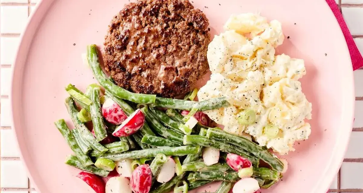 Un hamburger savoureux avec une salade de haricots verts et radis, et une salade de pommes de terre crémeuse sur une assiette rose.