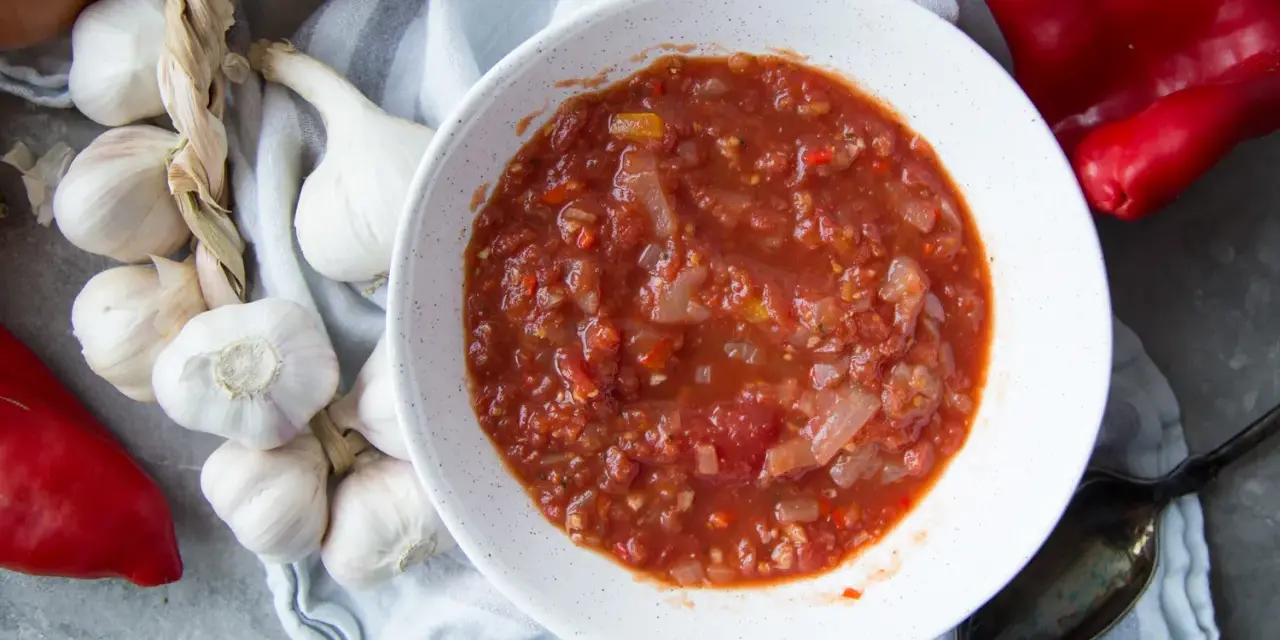 Un bol de salsa facile, avec des tomates, des oignons et des poivrons rouges. Des gousses d'ail et un oignon sont disposés autour.