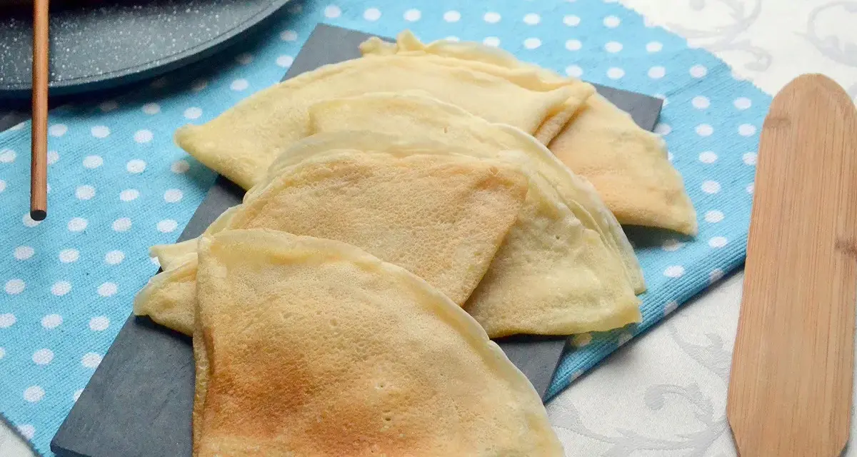 Pile de crêpes dorées, prêtes à être garnies. La pâte à crêpe vegan est légère et aérienne. Un ustensile en bois est à côté.