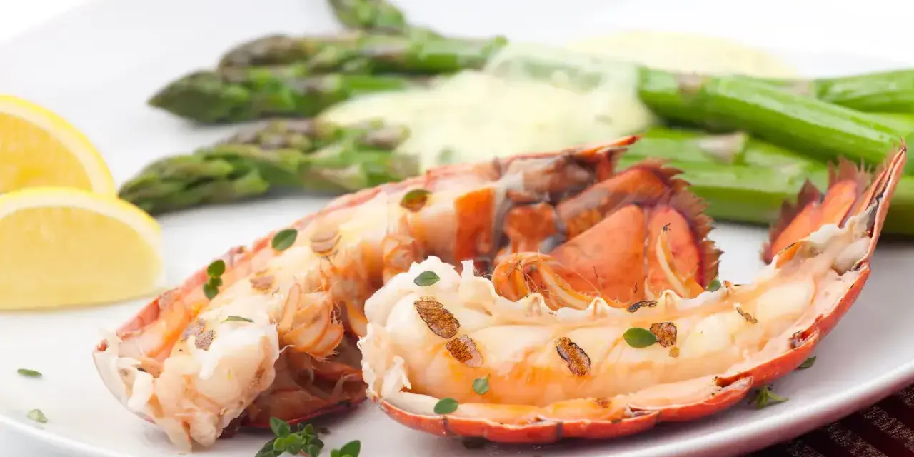 Queue de homard grillée, servie avec des asperges et une sauce homard crémeuse, garnie de quartiers de citron et de thym frais.