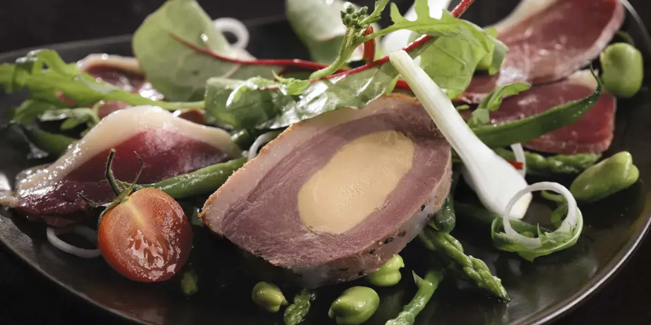 Une délicieuse salade landaise avec du foie gras, du magret de canard, des asperges et des tomates cerises sur une assiette noire.