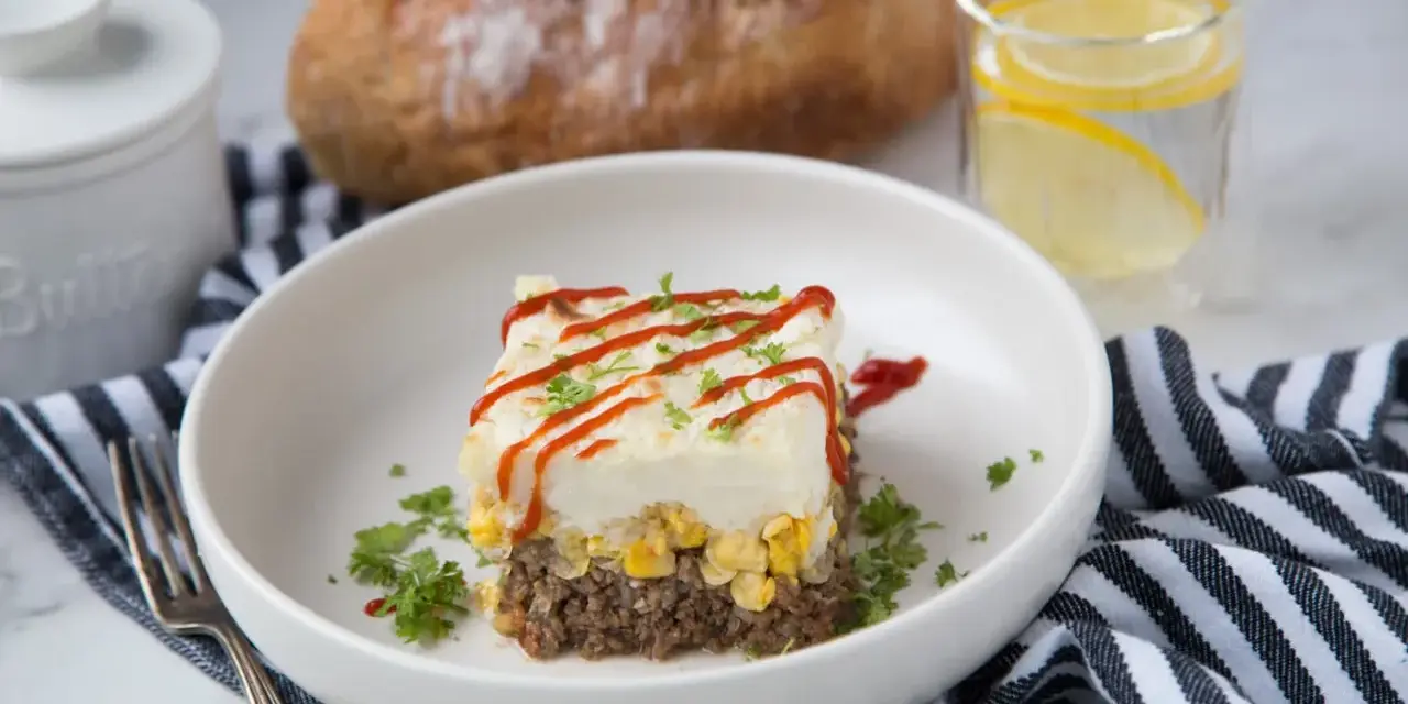 Un délicieux pâté chinois servi dans une assiette blanche, garni de persil et de ketchup. Un verre d'eau citronnée et du pain complètent ce plat réconfortant.