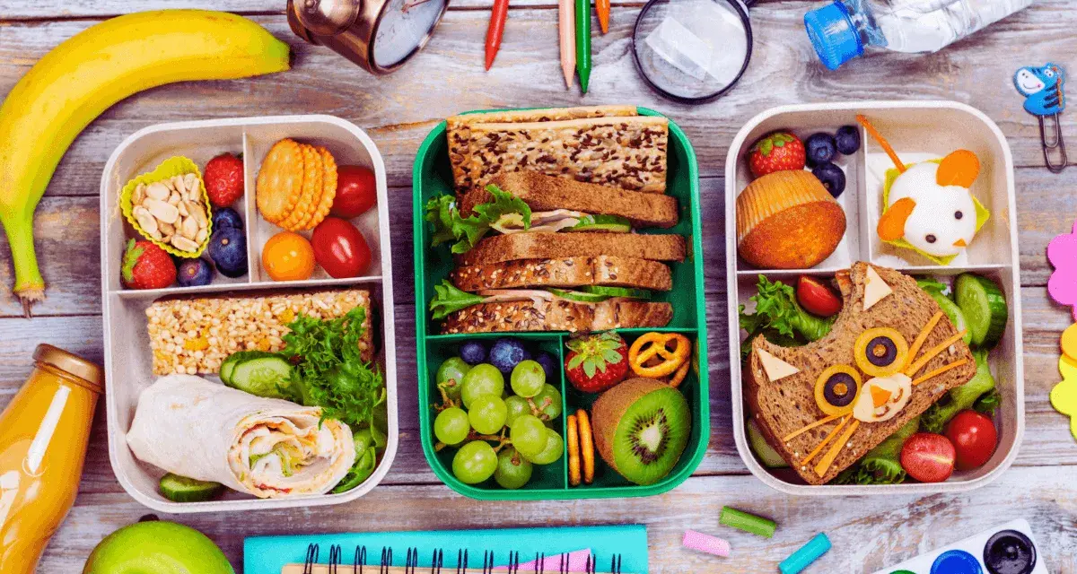 Trois boîtes repas à emporter faites maison, remplies de fruits, sandwichs créatifs et barres de céréales. Parfait pour un pique-nique ou un déjeuner sur le pouce.