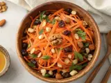 Salade de carottes râpées, garnie de noix, raisins secs et persil, dans un bol en bois. Un petit ramequin de miel est à côté.