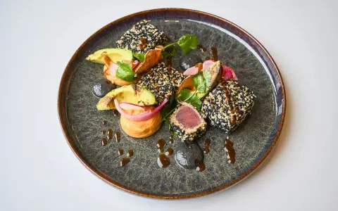 Un délicieux tataki de thon enrobé de sésame, servi avec de l'avocat, des oignons rouges et une sauce onctueuse sur une assiette texturée.